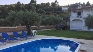 Une piscine à l’eau bleue, entourée d’une terrasse en béton, et quelques chaises longues. On aperçoit, au fond, un bâtiment blanc doté d’un balcon.