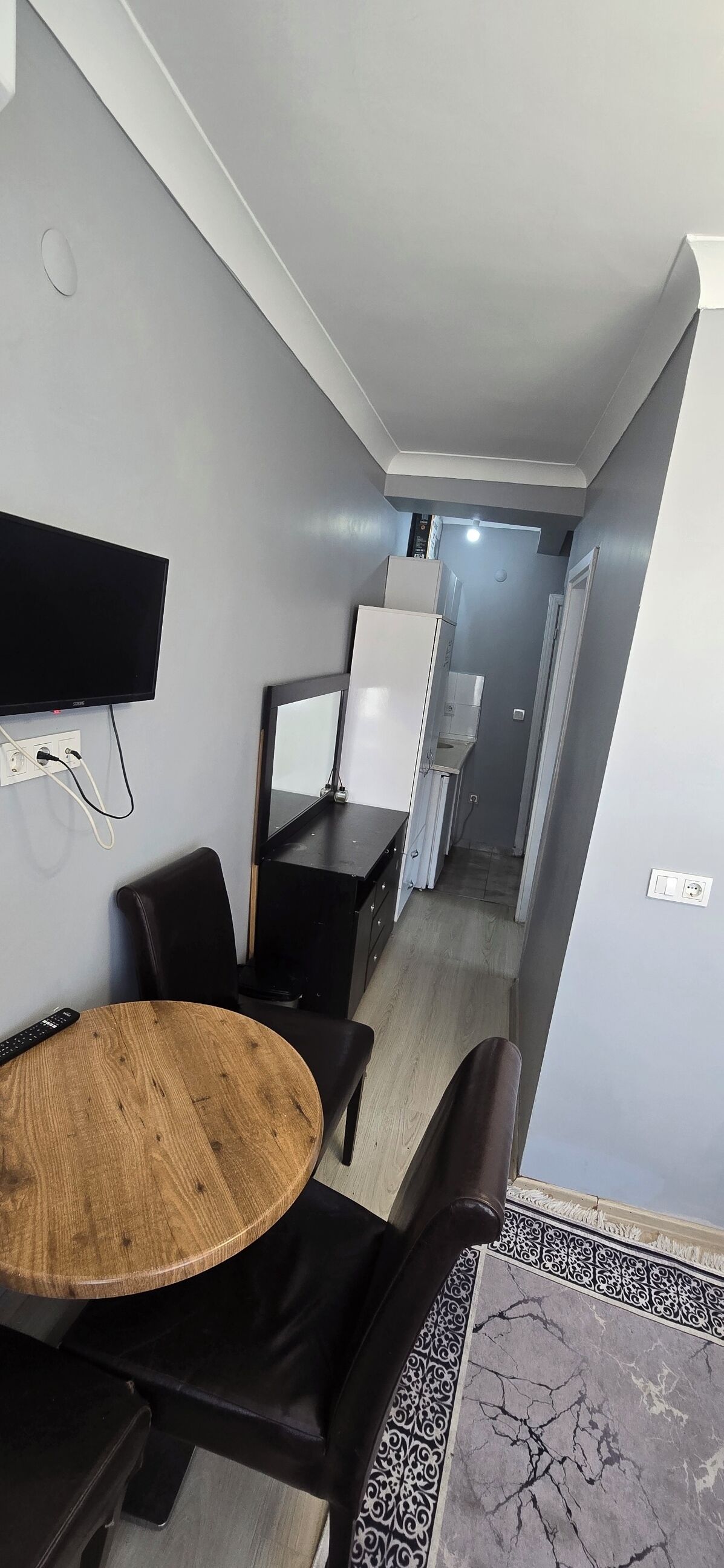 Un salon moderne avec une table ronde en bois, des chaises noires, un téléviseur à écran plat fixé au mur et un coin cuisine visible à travers une porte ouverte.