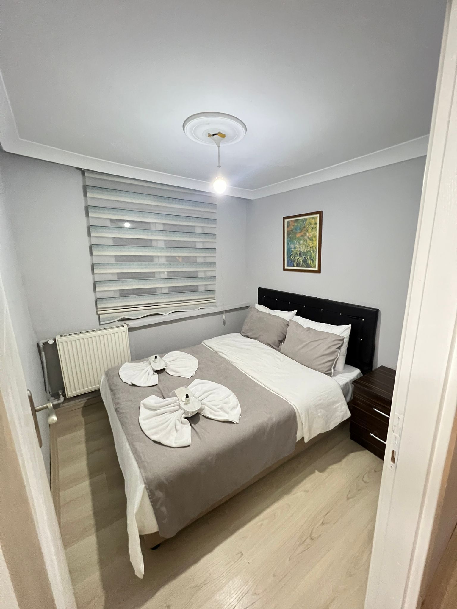 Quarto bem arrumado com uma cama, um radiador, janela com persianas, mesa de cabeceira e um quadro na parede.