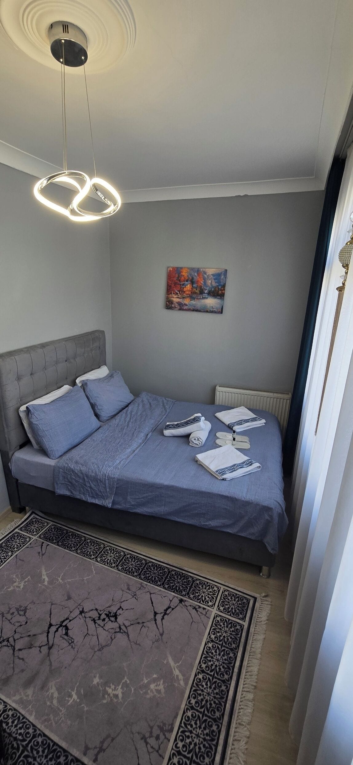 A bedroom with a grey bed, a grey headboard, a painting on the wall, and a patterned rug.