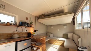 A compact kitchen with a refrigerator, stove, and wooden countertop. There is a small dining area with a bench and a window providing natural light. - The Secret Studio, Outdoor Bath & Pizza Oven (New Plymouth)