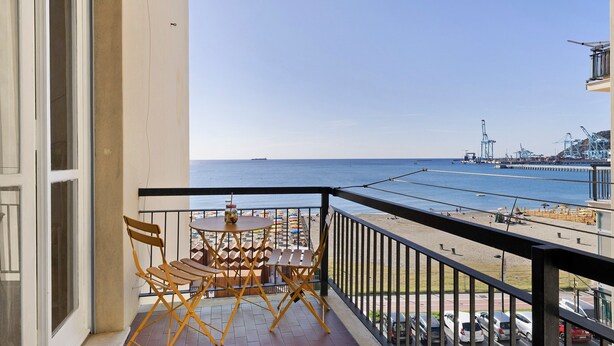 A balcony with a table and chairs, a view of the sea, and a distant port with cranes. - Holiday Apartment 'Riva Mare 2' Near the Beach with Sea View and Parking Space (Vado Ligure)