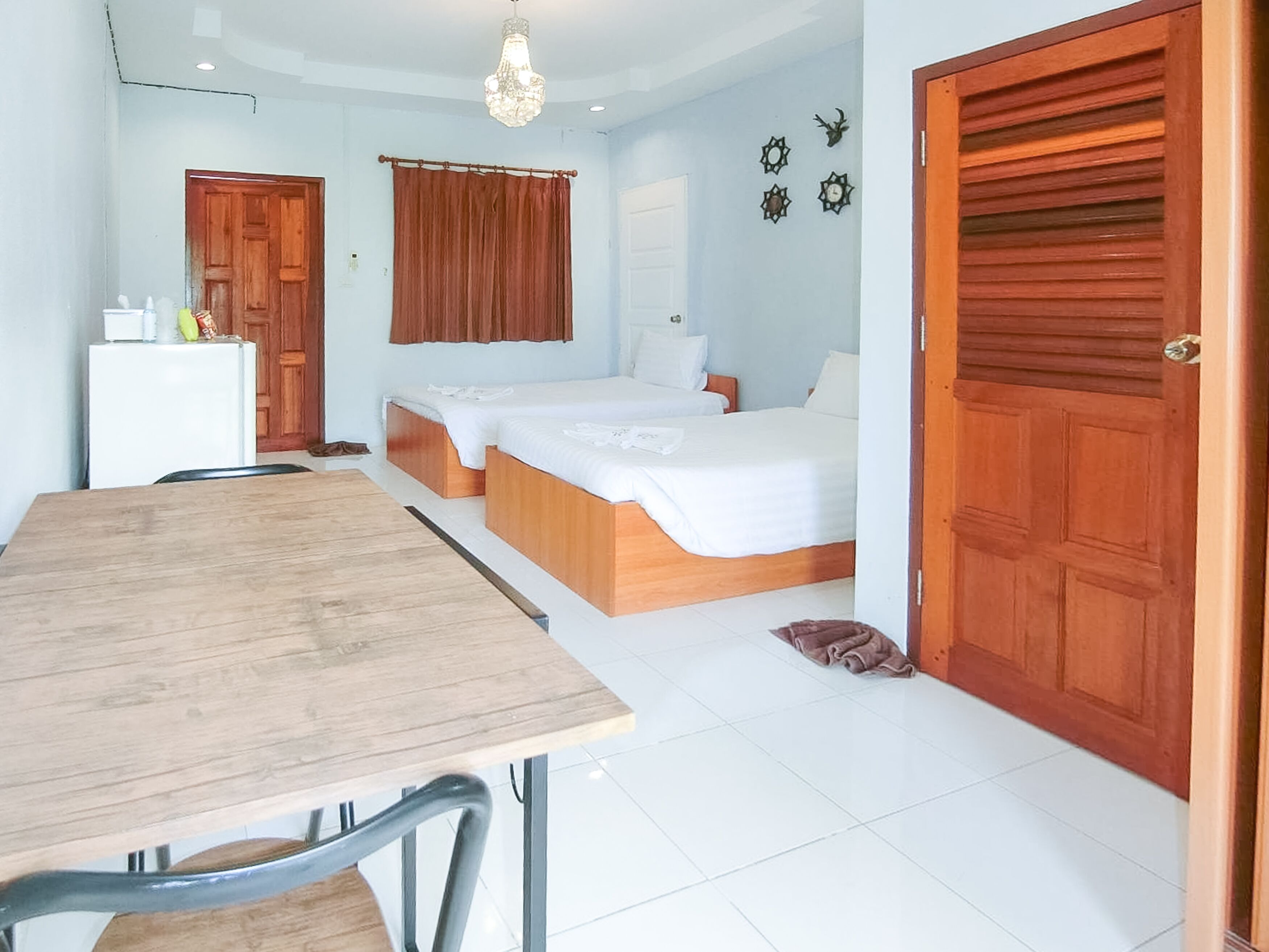 A room with a wooden table, two beds, a white wardrobe, and a door.