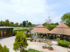 A courtyard with two thatched-roof structures, surrounded by greenery and a paved area with seating.