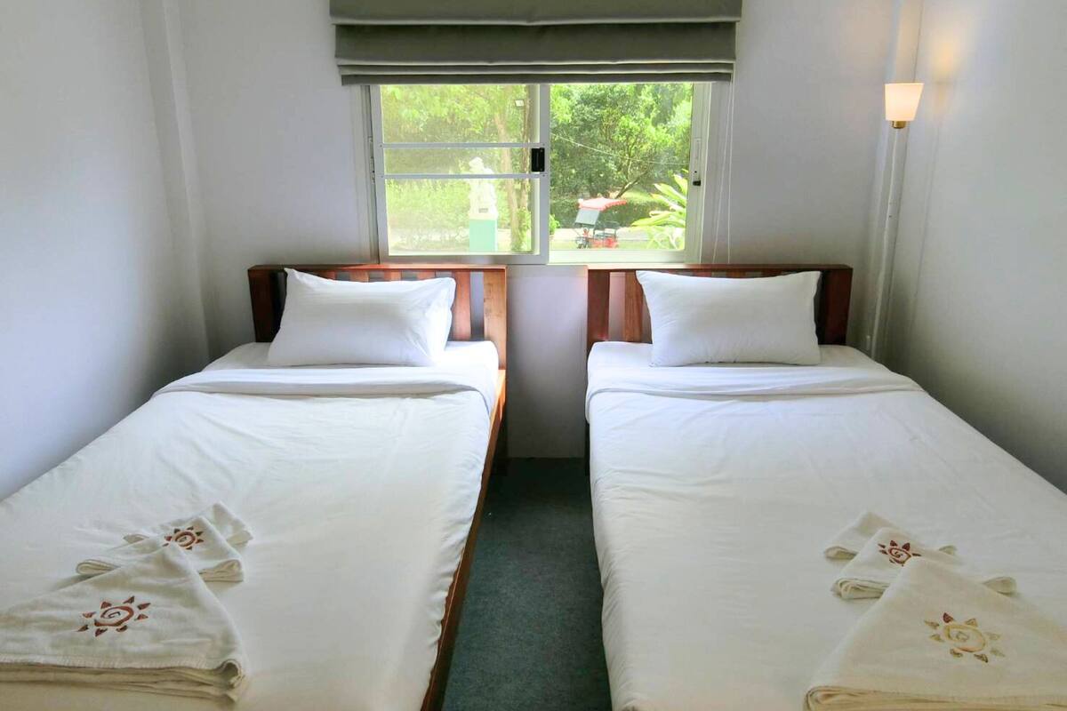 Ein Zimmer mit zwei Einzelbetten, jeweils mit weißer Bettwäsche und Kissen, einem Fenster mit Blick ins Grüne und einer Wandleuchte.