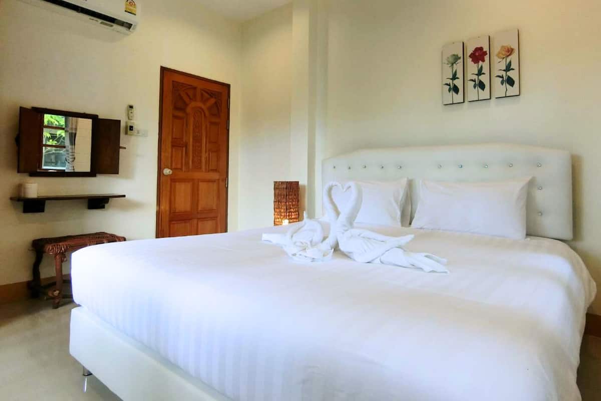 Ein Hotelzimmer mit einem großen Bett, einer Holztür, einer Wand-Klimaanlage, einer kleinen Ablage mit einem gerahmten Bild und einer Wand mit drei gerahmten Blumenbildern.