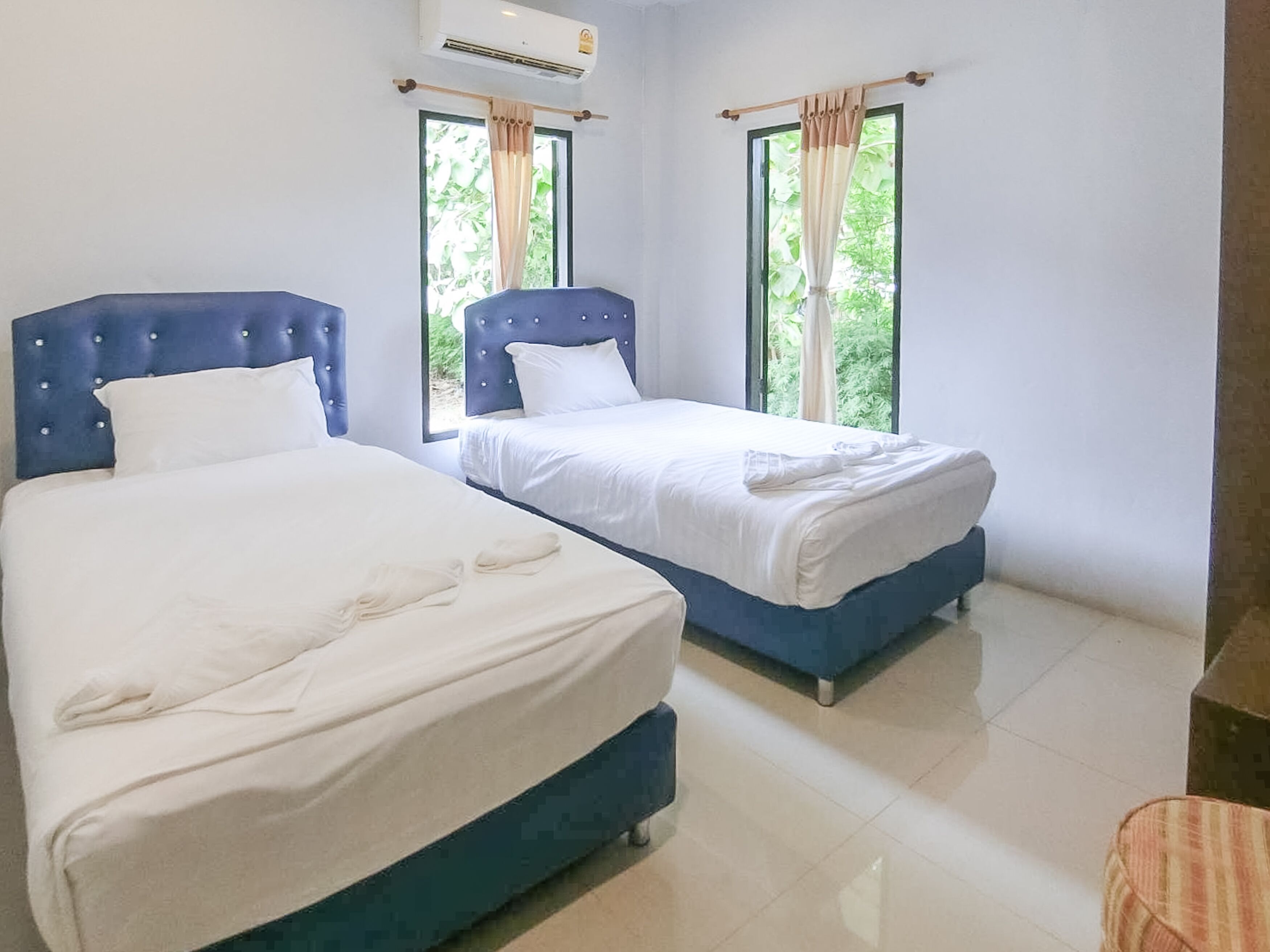 A hotel room with two beds, each with a blue headboard and white bedding. There are windows with curtains, and an air conditioning unit is visible on the wall.