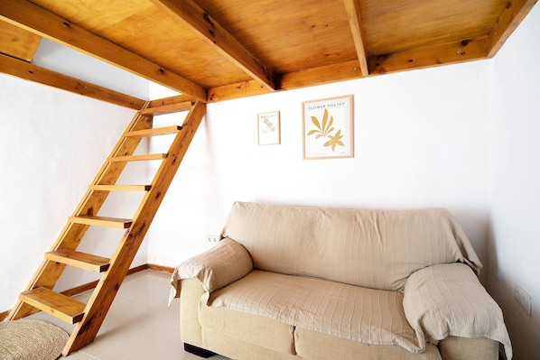 Une échelle en bois adossée à un mur blanc, avec deux œuvres d’art encadrées, un canapé beige et un tapis tissé.
