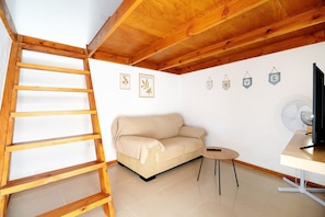 Une pièce avec une échelle en bois, un canapé beige, une petite table ronde, un téléviseur et des éléments décoratifs sur les murs.