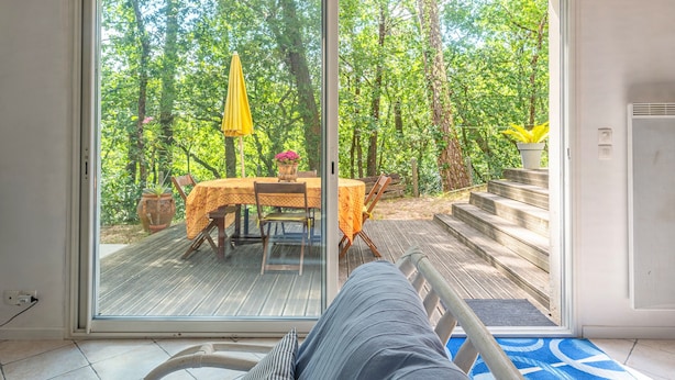Een kamer met een schuifdeur die uitkomt op een terras met een tafel en stoelen, omgeven door bomen.