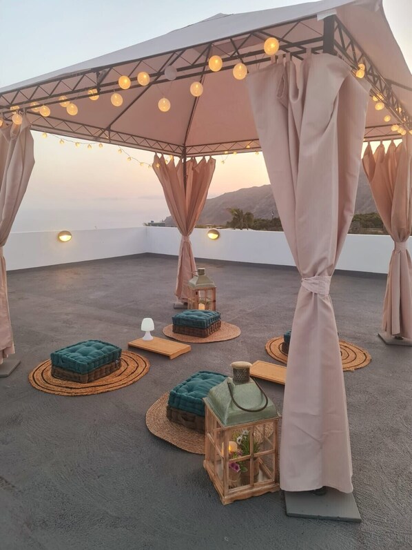 A rooftop terrace with a canopy, string lights, cushioned seating, and a lantern.