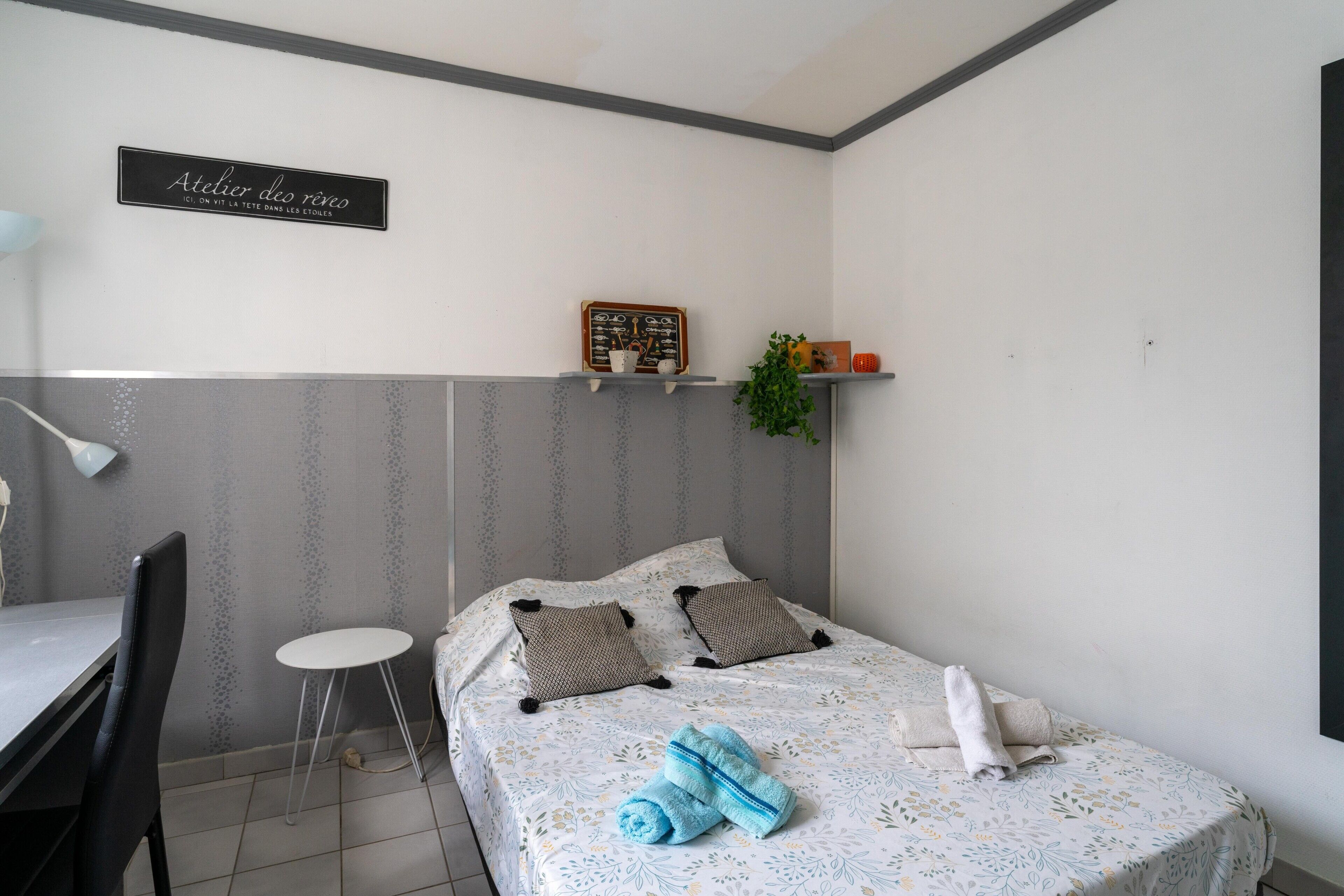 Un dormitorio con cama, una mesita, una silla, una estantería con una planta y una pizarra en la pared.