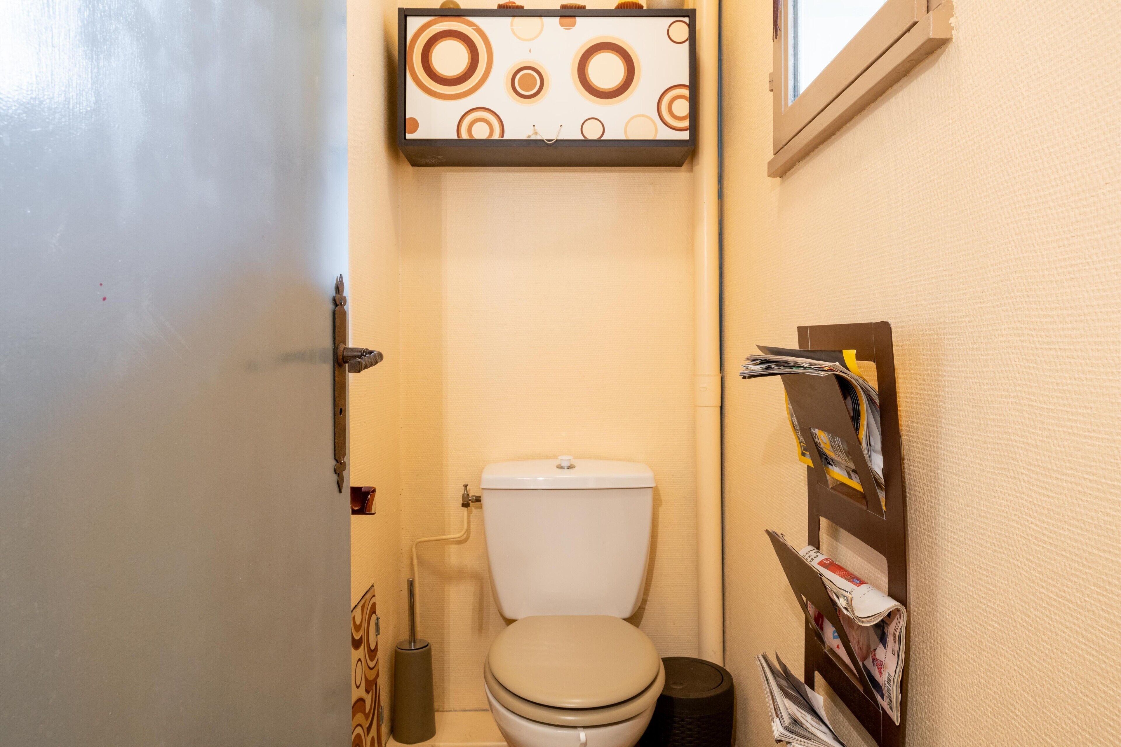 Une petite salle de bain avec des toilettes, une étagère fixée au mur où sont posés des magazines et une fenêtre.