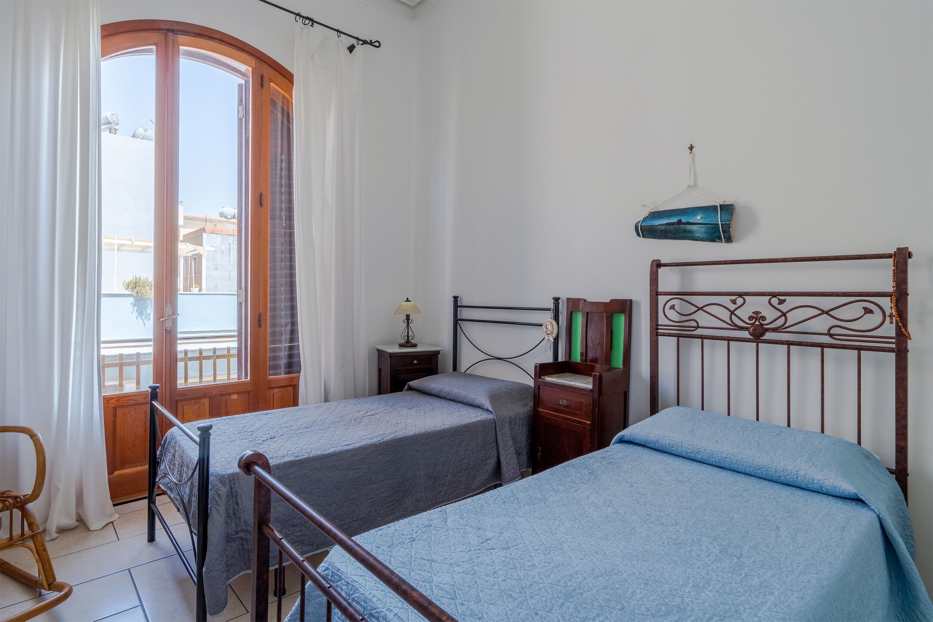 A room with two beds, a wooden door, a small table with a lamp, and a hanging item on the wall.