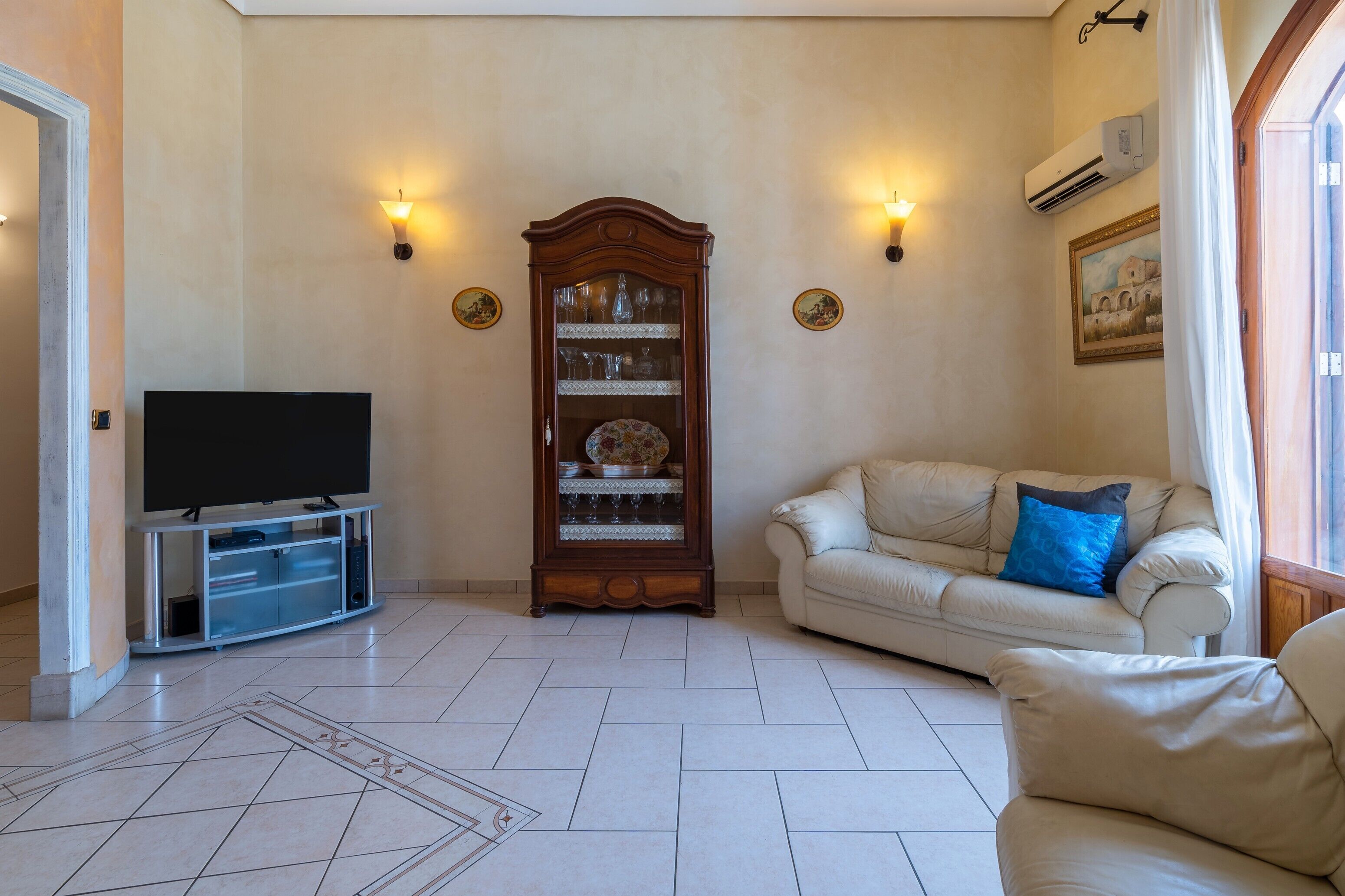 A living room with a flat-screen TV, a wooden cabinet with glass doors, a beige sofa with a blue pillow, and wall-mounted lights.