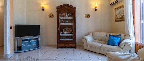 A living room with a flat-screen TV, a wooden cabinet with glass doors, a beige sofa with a blue pillow, and wall-mounted lights.