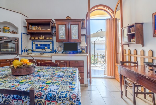 Une cuisine avec un mur recouvert de carreaux, des placards en bois, une table à manger avec une nappe à motifs et une vue sur l’extérieur.
