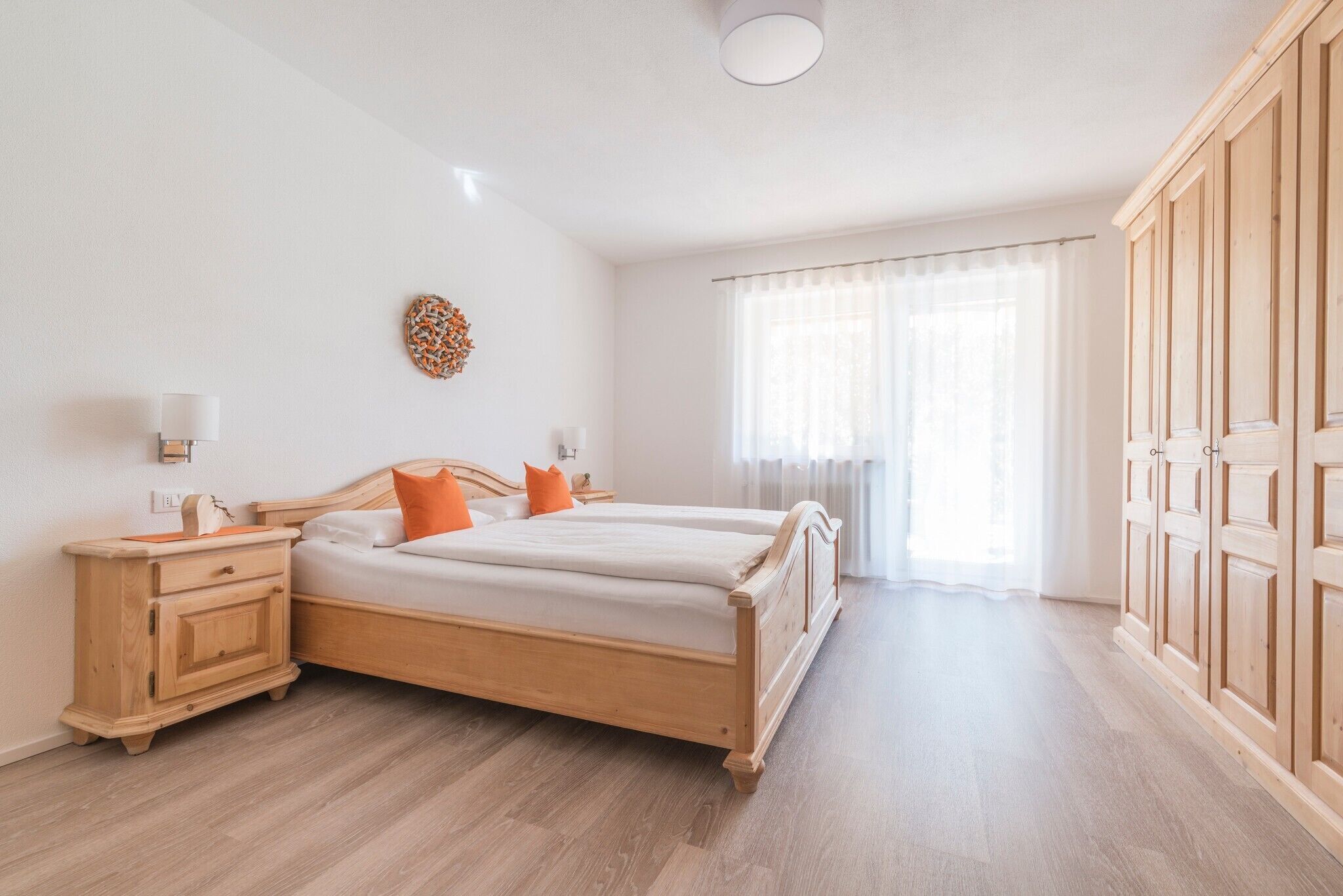 A bedroom with a wooden bed, a nightstand, a wardrobe, and a window with sheer curtains.