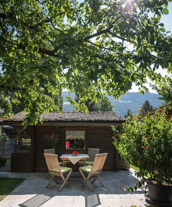 A wooden outdoor seating area with a table and chairs, surrounded by greenery and a view of the mountains.