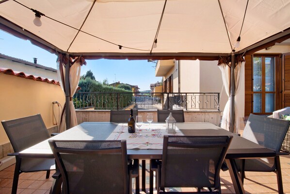 A covered outdoor dining area with a table set for two, chairs, a bottle, and a glass.