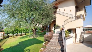 A building with a stone pathway leading to a gated entrance, surrounded by greenery and a large tree.