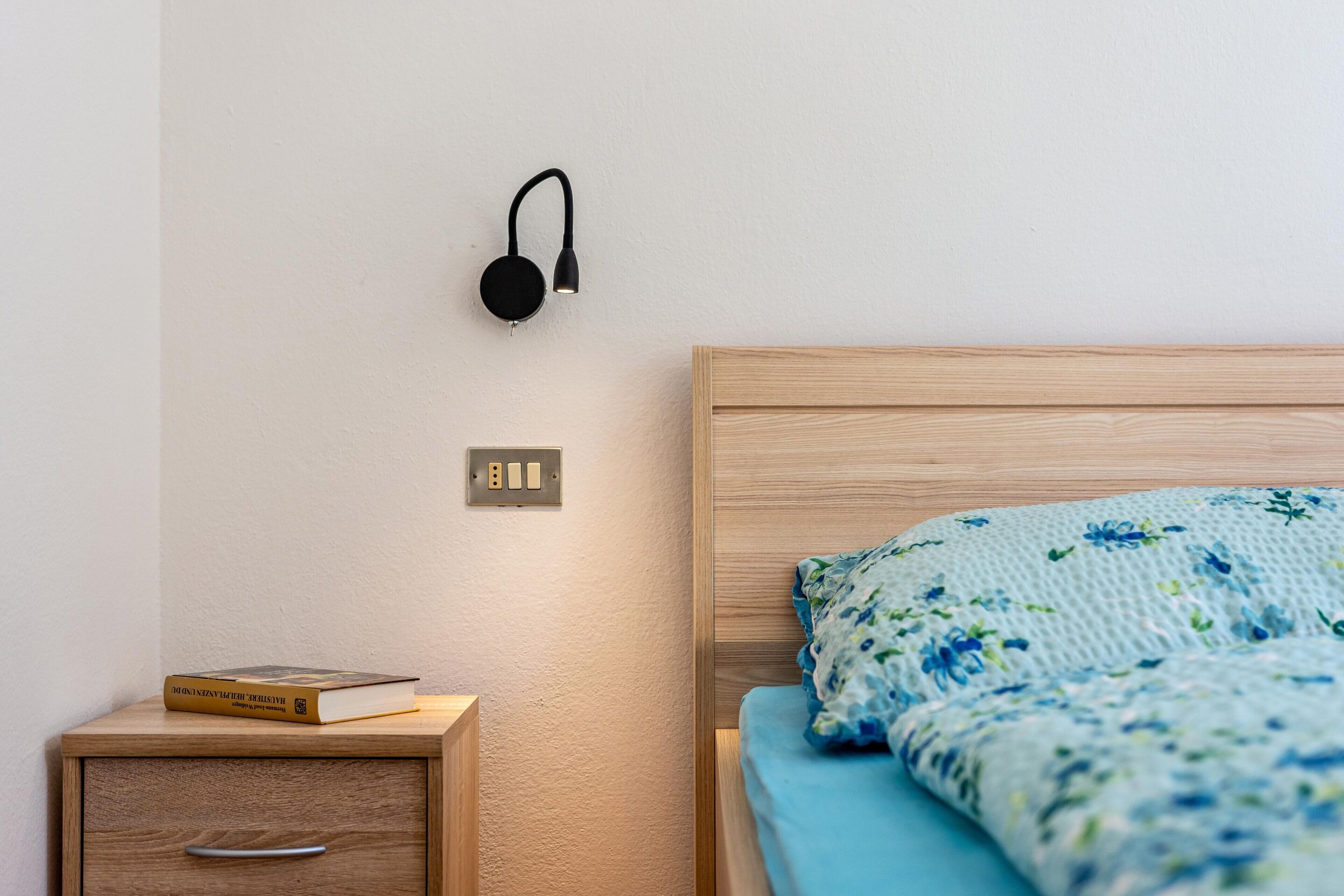 A neatly made bed with a floral-patterned bedspread, a bedside table with a book, and a wall-mounted lamp.