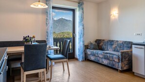 A room with a dining table, chairs, a sofa, a view of mountains, and a balcony with curtains.