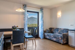 A room with a dining table, chairs, a sofa, a view of mountains, and a balcony with curtains.