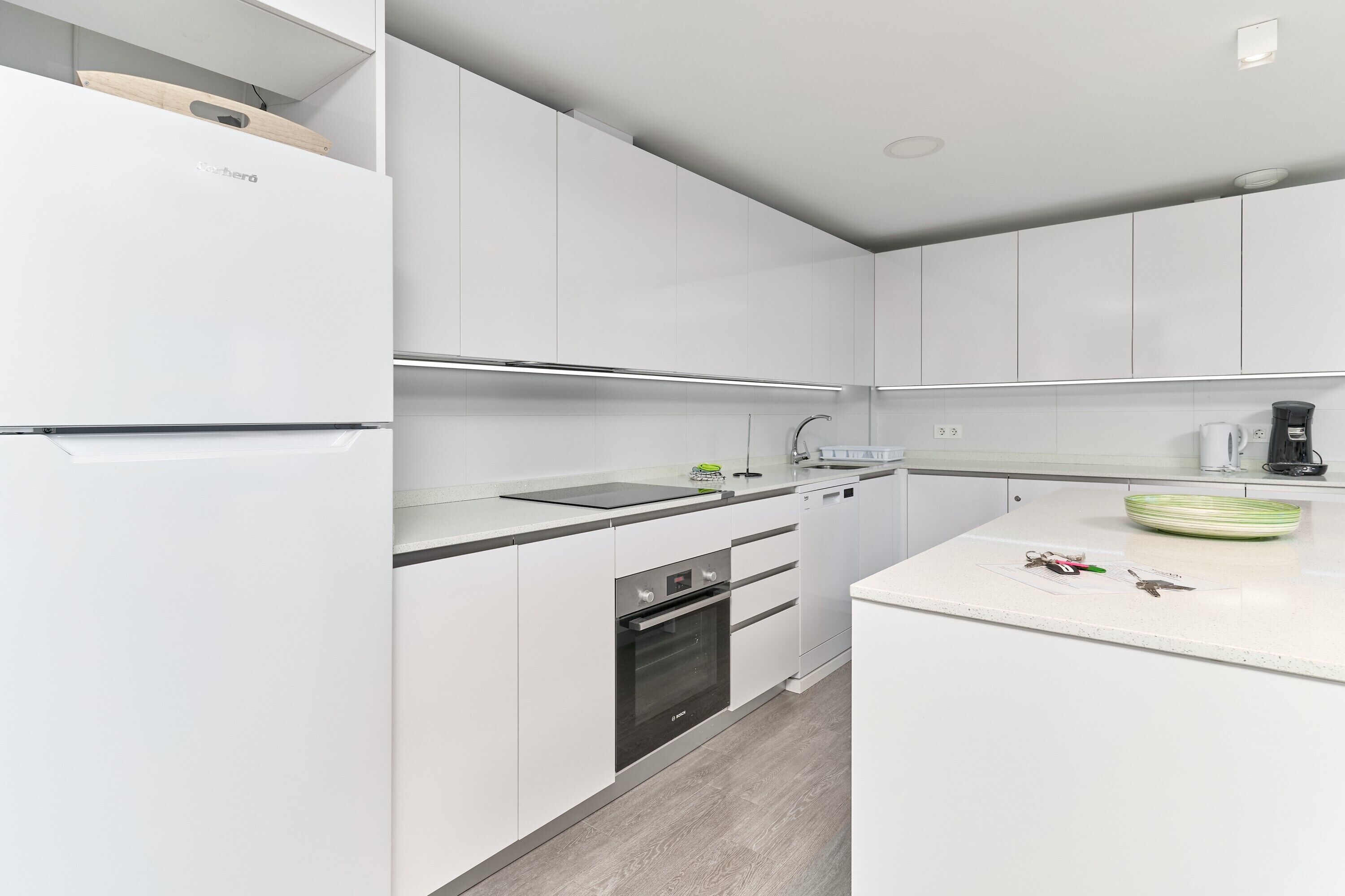 Una cocina moderna con armarios blancos, horno integrado y encimera con cafetera y un bol.