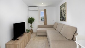 Ein modernes Wohnzimmer mit Flachbildfernseher, einem beigefarbenen Sofa, einem kleinen Topfpflanzen und einem Wandbild.