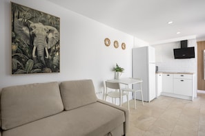 A modern living room with a beige sofa, a dining area with a white table and chairs, a kitchen with white cabinets, and a mounted TV.