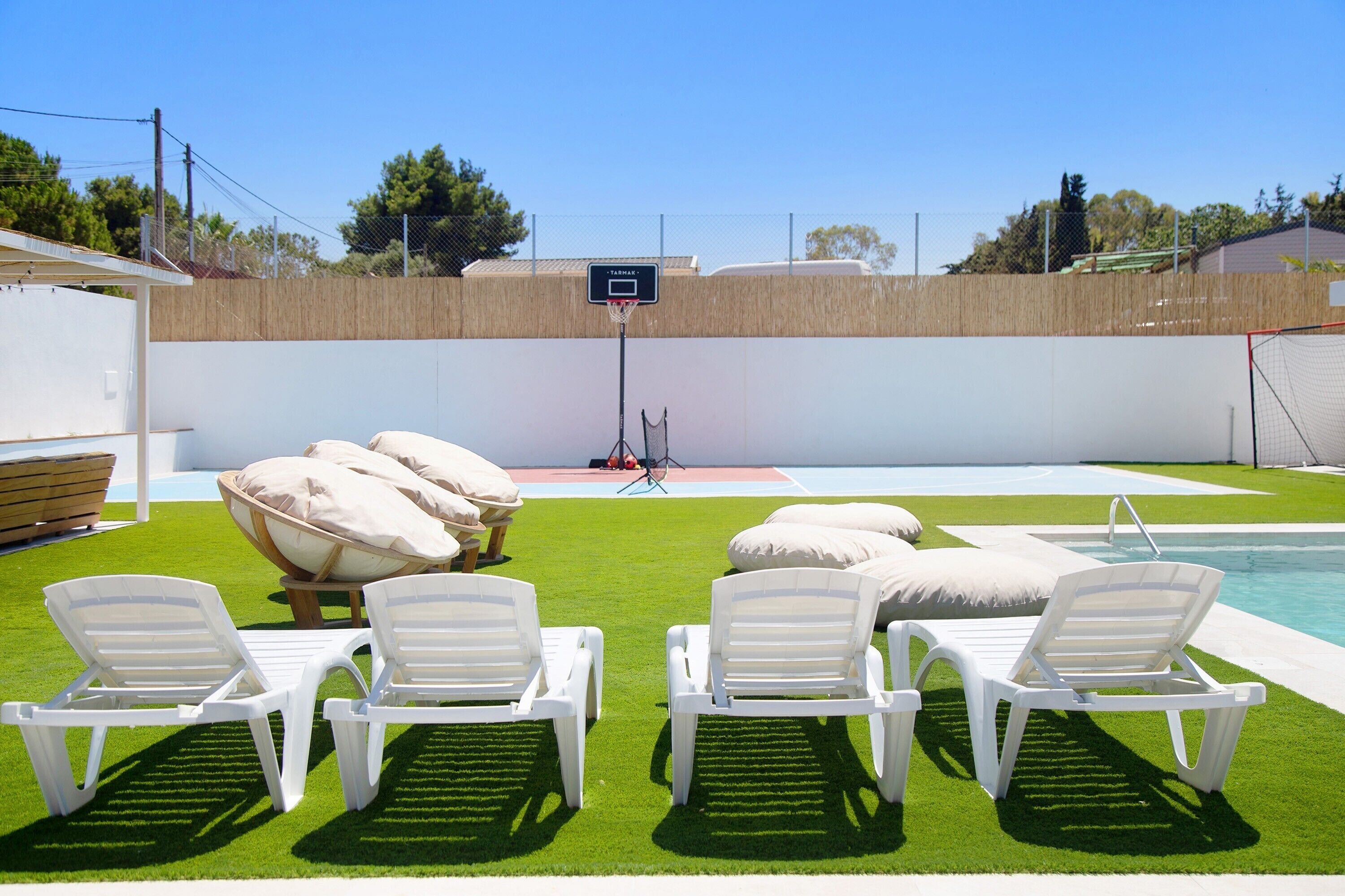 Zona de piscina con césped artificial, sillas de descanso blancas y aro de baloncesto.