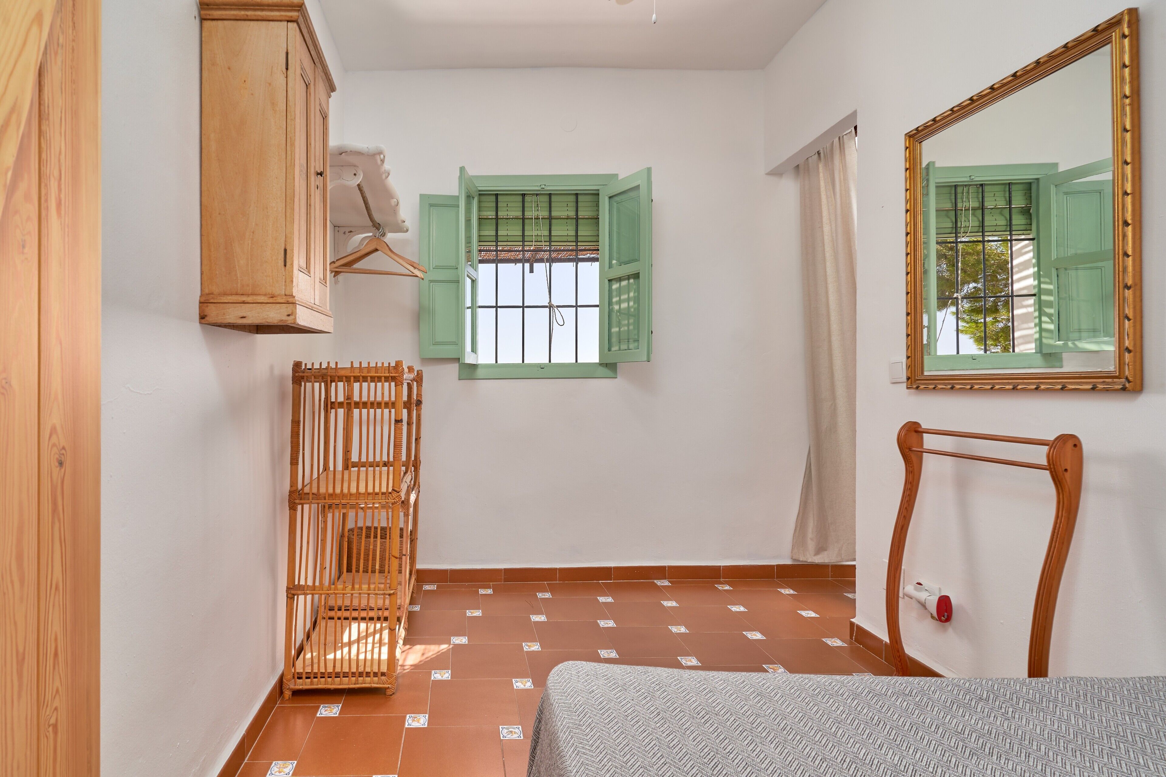 Ein Zimmer mit einem Bett, einem Holzschrank, einem grünen Fenster, einem Spiegel und einem gefliesten Boden.