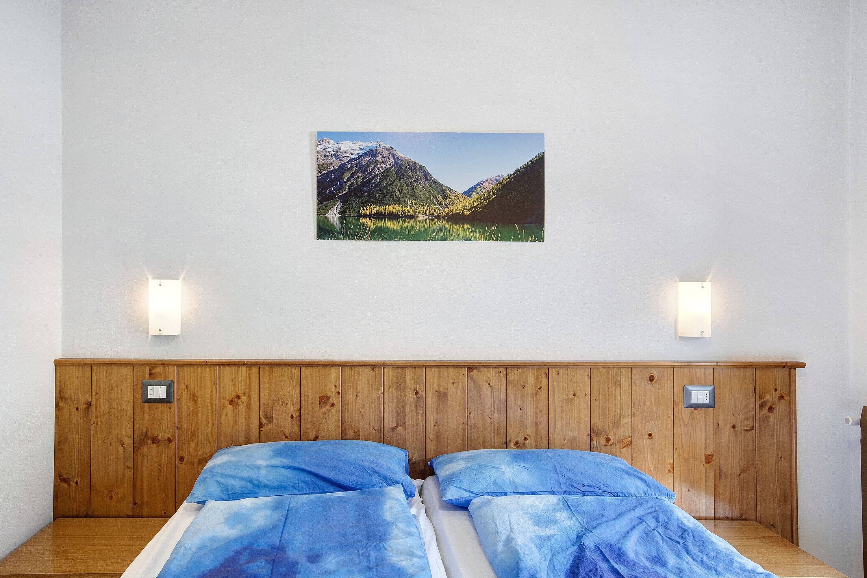 Ein Zimmer mit einem hölzernen Kopfteil, zwei Betten mit blauer Bettwäsche, einem an der Wand hängenden Bild von einer Berglandschaft und zwei Wandleuchten.