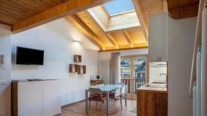 A modern kitchen with a skylight, wooden ceiling beams, a dining area with chairs, and a TV mounted on the wall.