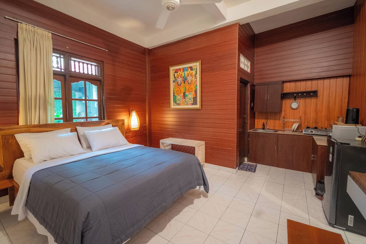 A bedroom with a bed, a nightstand, a desk, a window with curtains, a wall-mounted painting, and a kitchenette.