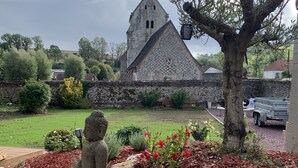 A stone church with a tall spire, surrounded by a landscaped garden with red mulch, rocks, and various shrubs. - Villa Marguerite, à 12 Minutes de la Plage ! (Cormont)