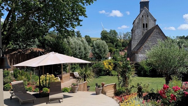 A covered outdoor area with a wooden deck, lounge chairs, and a pergola with an umbrella. In the background, there is a church with a tall spire and wind turbines. - Villa Marguerite, à 12 Minutes de la Plage ! (Cormont)