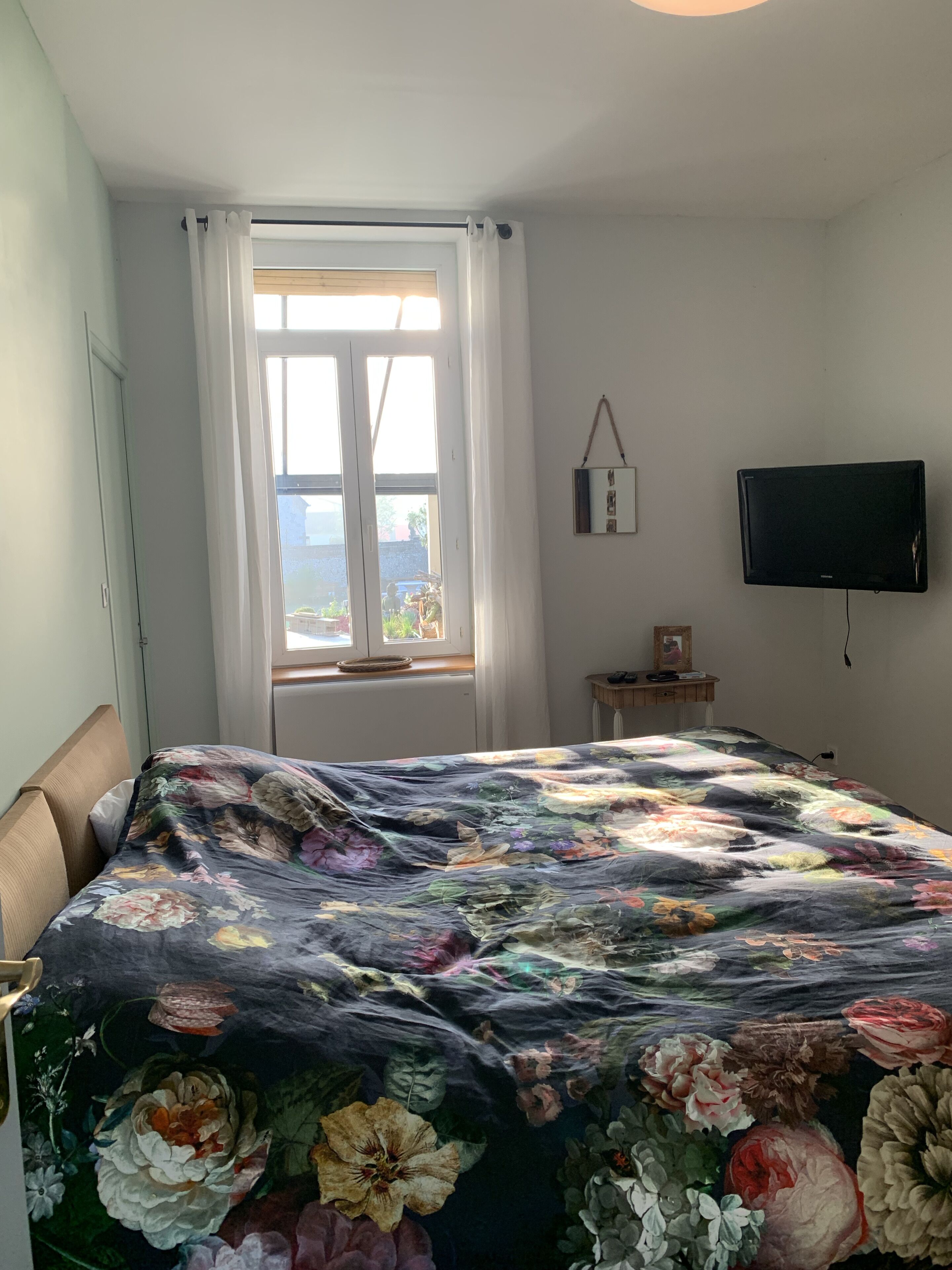 A bedroom with a large floral bedspread, a window with curtains, a bedside table with a lamp, and a mounted TV.