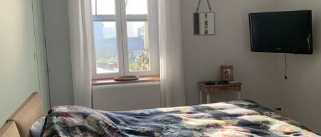 A bedroom with a large floral bedspread, a window with curtains, a bedside table with a lamp, and a mounted TV.