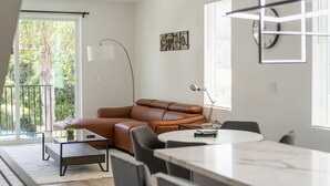 Een moderne woonkamer met een bruine leren bank, een glazen salontafel en een eethoek met een marmeren tafel.