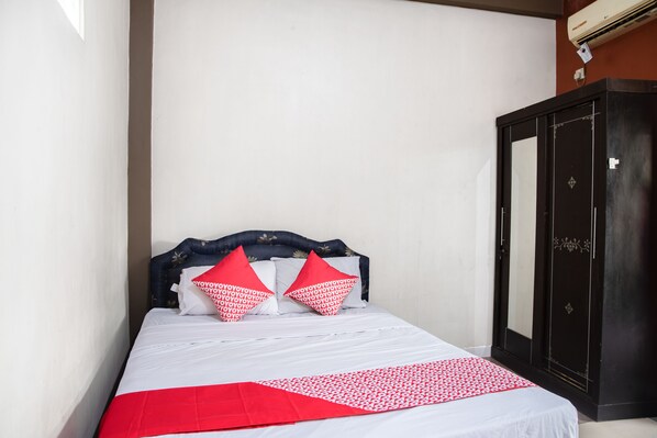Una cama bien hecha con ropa de cama blanca y detalles rojos, un armario de madera oscura y una pared lisa.