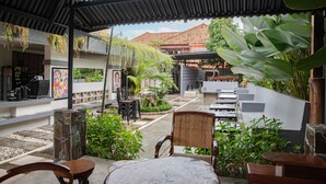 An outdoor patio area with stone pillars, wooden chairs, and tables. There are potted plants and a view of a building with a tiled roof in the background.
