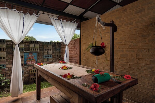 Una mesa de madera con mantel, rodeada de flores, en un espacio exterior cubierto con paredes de ladrillo y un mural.