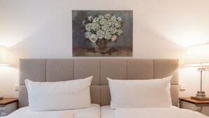 Habitación de hotel con una cama, dos almohadas blancas, un cabecero, un cuadro de flores y dos lámparas de noche.