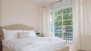 Una cama bien hecha con sábanas y almohadas blancas, una mesita de noche con lámpara, y un ventanal con cortinas.