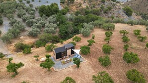 Una pequeña cabaña moderna con terraza y asientos exteriores, rodeada de olivos y terreno árido.