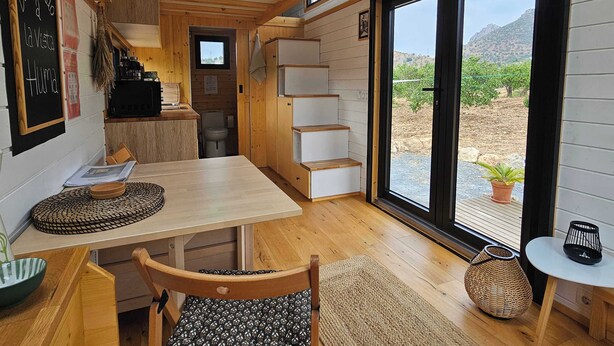 Un interior acogedor con una mesa de madera, sillas, una pequeña cocina y un ventanal con vistas al exterior.