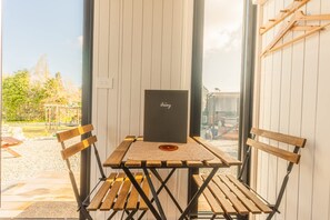 Une table en bois sur laquelle se trouve un objet noir, entourée de chaises en bois, avec vue sur un jardin et un bâtiment en arrière-plan.