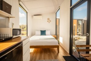 Une chambre moderne avec un lit en bois, une table de chevet, un four à micro-ondes encastré et une grande fenêtre donnant sur l’extérieur.